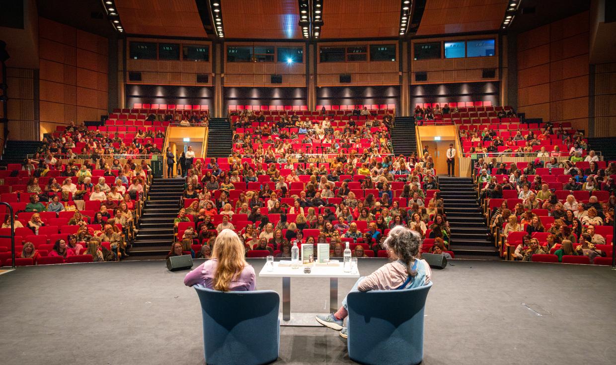 A shot from the back of the auditorium stage, looking at the back of Caitlin and Moran and host, as they look out to the large audience.